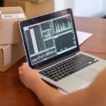 Osoba analizująca dane na laptopie obok paczek w małym sklepie internetowym